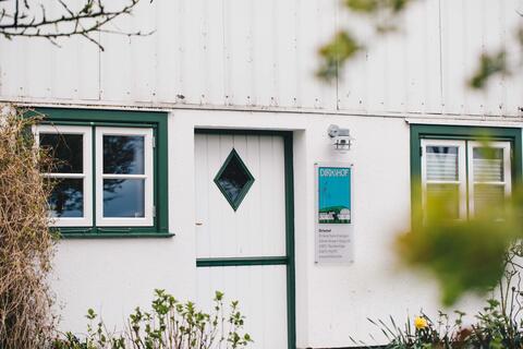Der Eingang zum Hauptgebäude des Dirkshofs: Eine weiße, grün gerahmte Tür neben einem weißen, grün gerahmten Fenster. Neben der Tür hängt ein Schild mit dem Dirkshof-Logo.